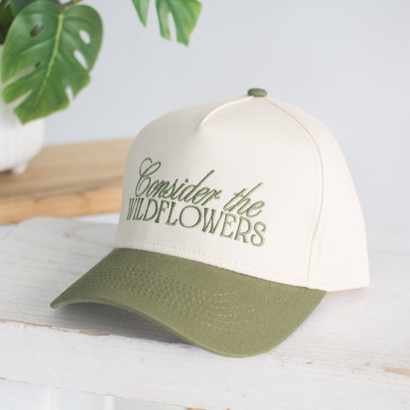 White cap with green brim and 'Consider the Wildflowers' text on a white surface with a plant in the background