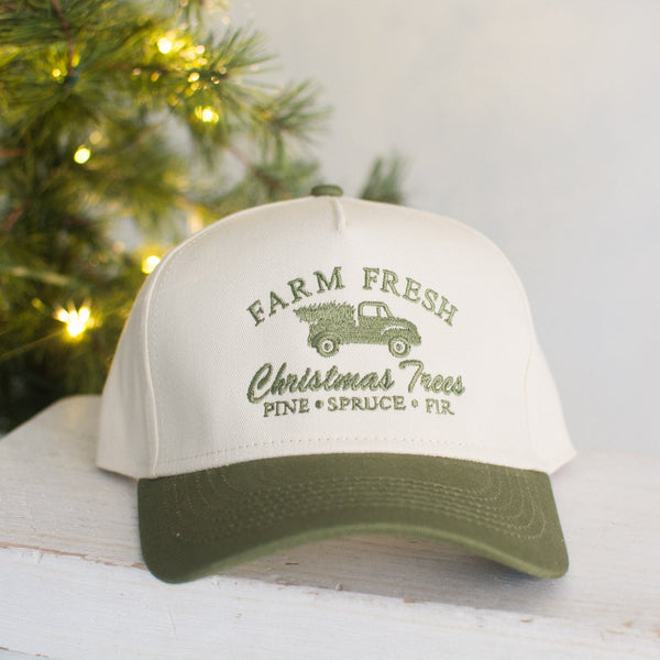 White cap with green brim featuring 'Farm Fresh Christmas Trees' design on a white surface with a Christmas tree in the background.