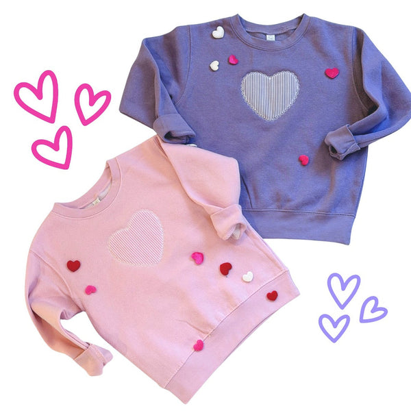 Two children's sweatshirts, one pink and one blue, with heart designs on a white background.